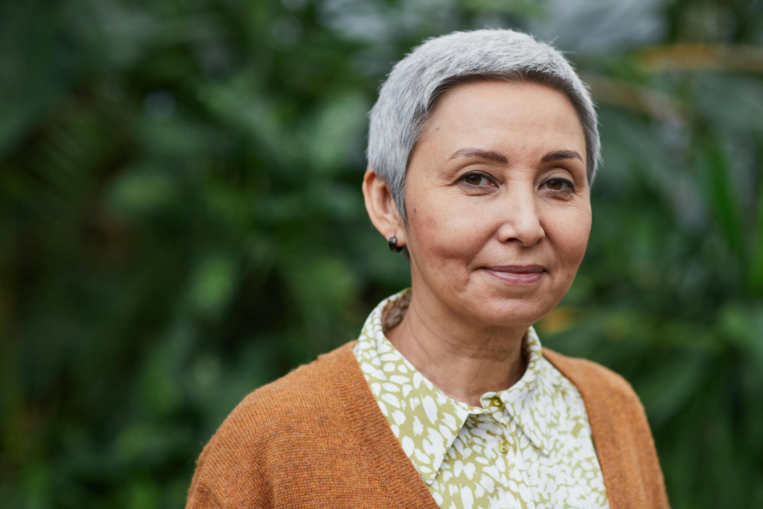 Portrait of an elegant senior Asian woman with a joyful expression, standing outdoors with greenery in the background.